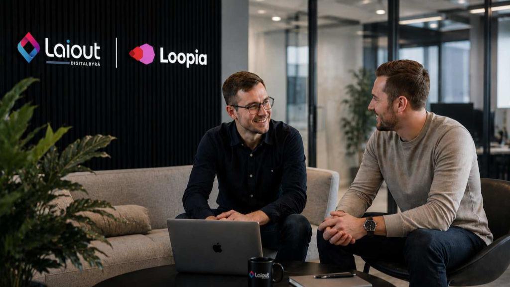 Två personer sitter i en modern kontorsmiljö och samtalar vid ett bord med laptop och anteckningsblock. I bakgrunden syns Laiout Digitalbyrås och Loopias logotyper i en stilren och professionell miljö som förmedlar samarbete och partnerskap som Loopia partner inom digital utveckling och webb.